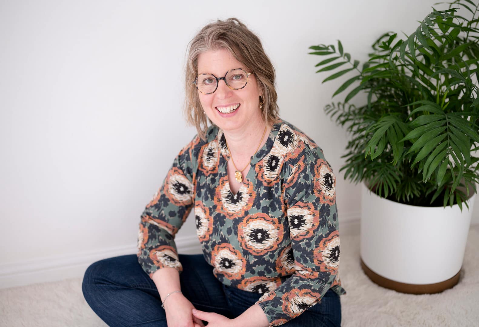 Gina sits on a neutral coloured floor in front of a green plant